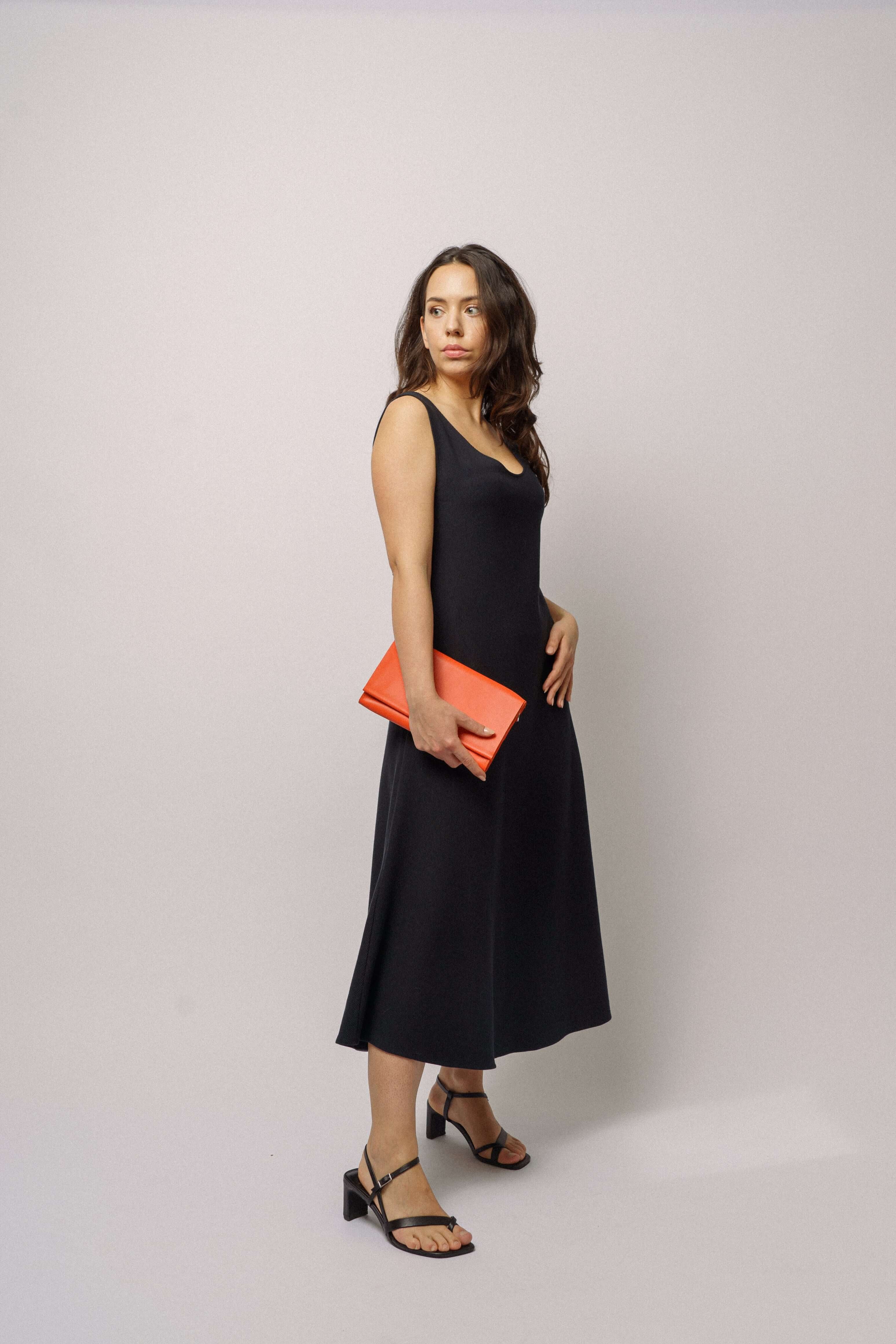 Modelka v černých dlouhých šatech bez rukávů ADVA Studios z organické bavlny s černými sandály a červenou kabelkou. 