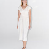 Modelka v bílých řasených šatech ADVA Studios s černými sandály na bílém pozadí. 