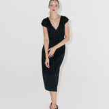 Modelka v černých řasených šatech s černými sandály na bílém pozadí. 
