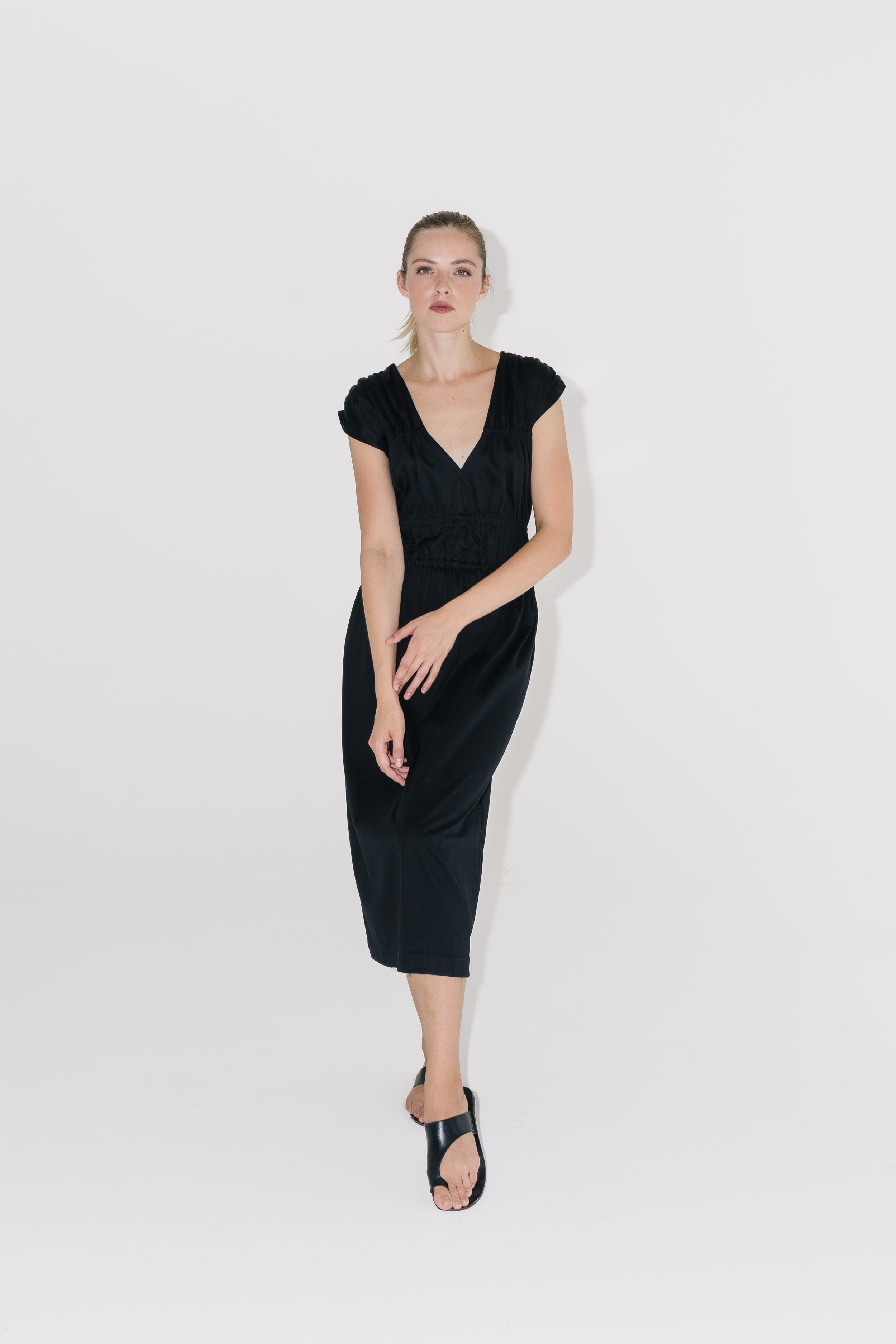 Modelka v černých řasených šatech s černými sandály na bílém pozadí. 