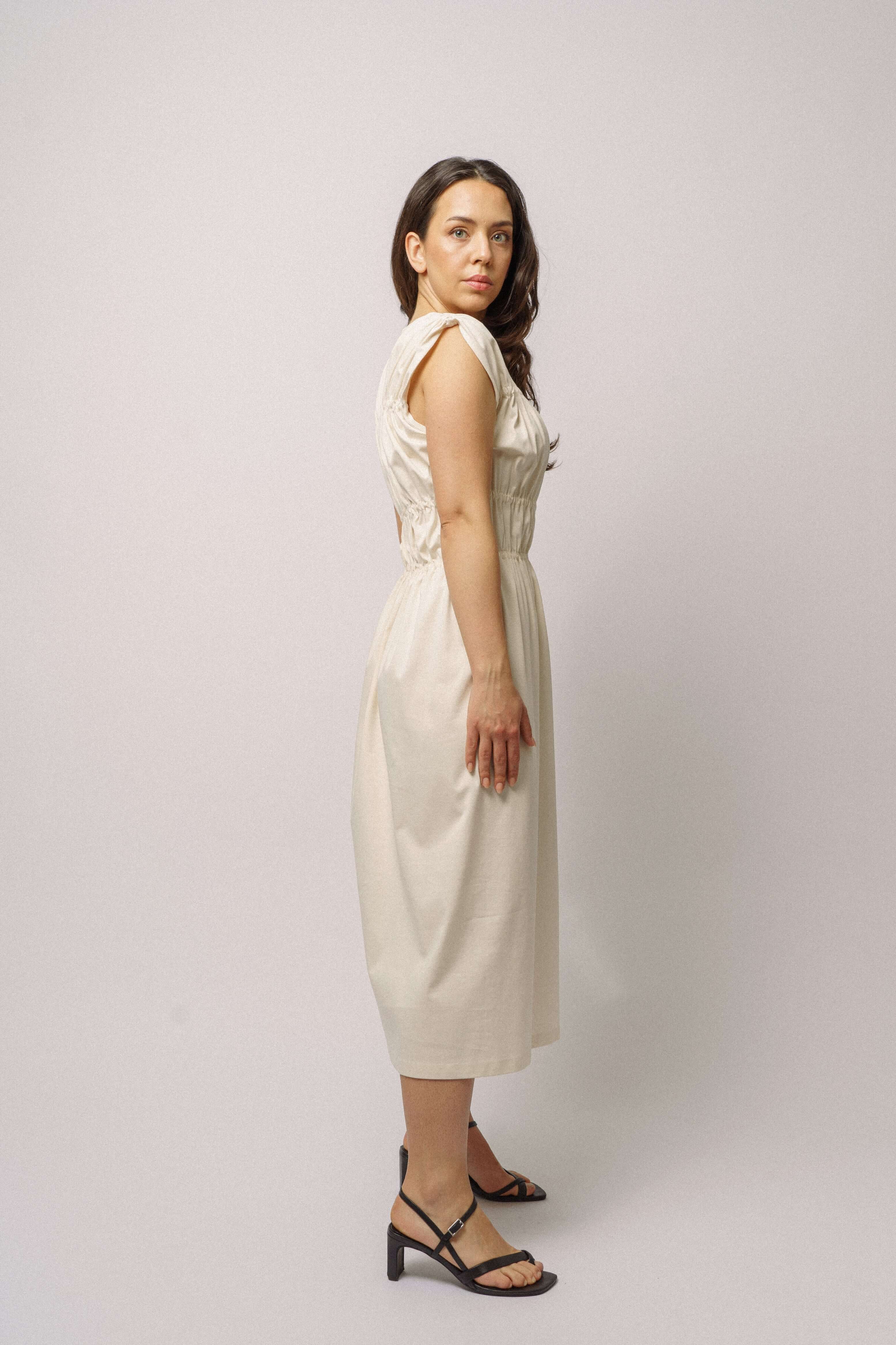 Modelka v bílých řasených šatech ADVA Studios s černými sandály na bílém pozadí. 